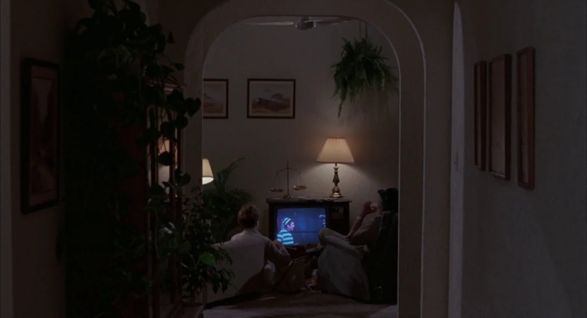Creepshow 2 (1987) movie still: wide — A dimly lit room viewed through an arched doorway from a dark hallway. Two people sit in armchairs facing a…