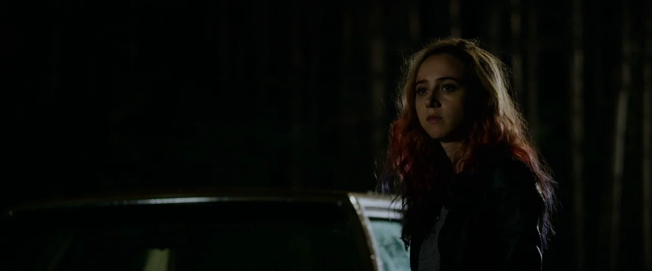 The Monster (2016) movie still: medium — A young woman stands near a vehicle at night in a dark wooded area. Her face is illuminated by a subtle…