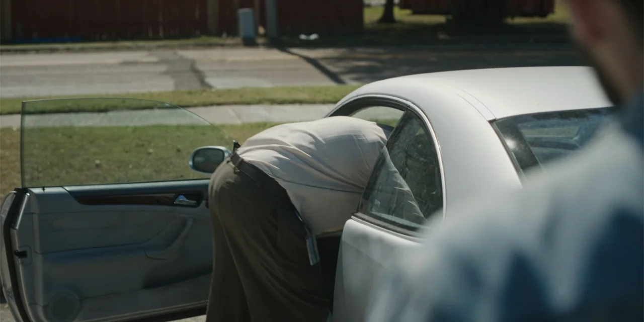 Caddo Lake (2024) movie still: medium — A silver car is parked on the side of a paved road with grass. A man in a beige shirt leans through the open…