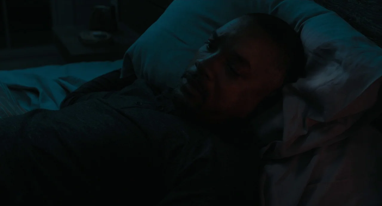 Gemini Man (2019) movie still: closeup — A dark, dimly lit bedroom at night with soft blue light hitting the bed linens. A man with short hair lies…
