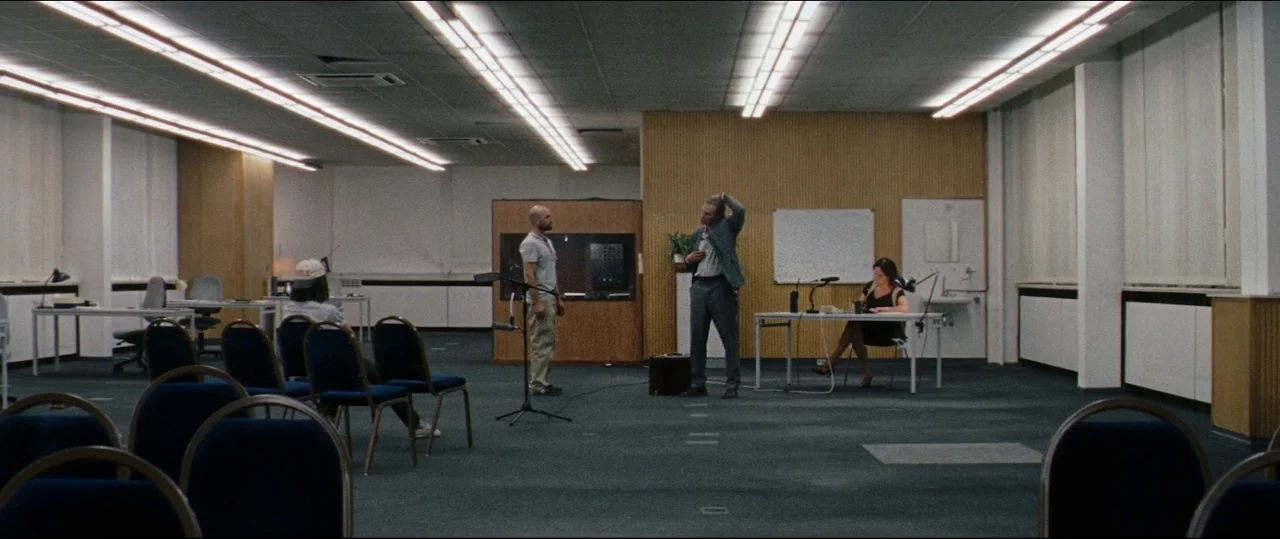 Luz (2018) movie still: wide — A spacious room features rows of blue fabric chairs and long white tables. Three people interact in the center of the…