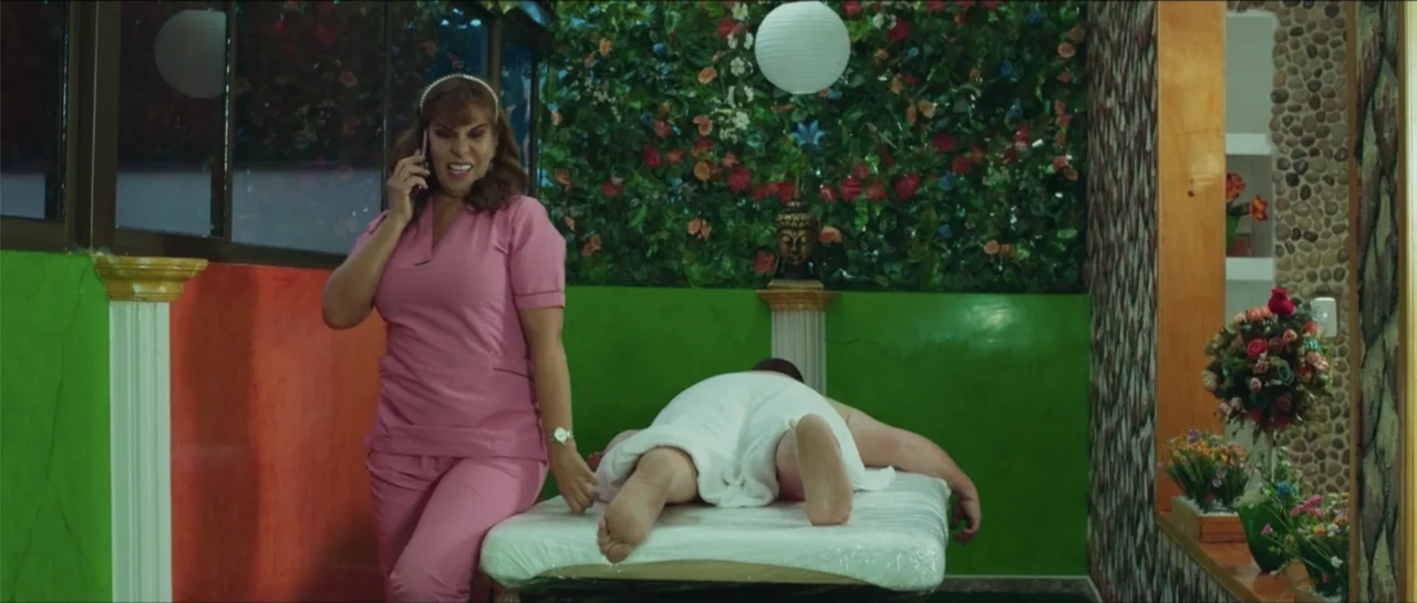 Loving is Losing (2019) movie still: medium — A massage room features bright green walls decorated with faux flowers and a Buddha bust. A woman in…