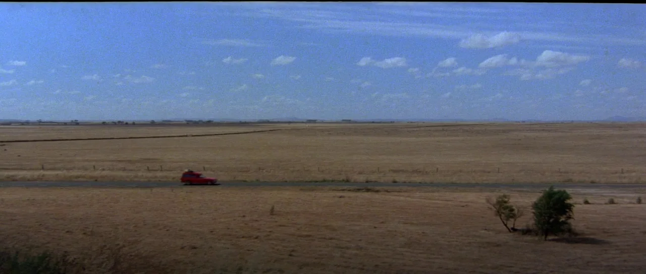 Mad Max (1979) movie still: establishing — A vast, flat expanse of dry, golden-brown grass stretches to the distant horizon line. A small red car…