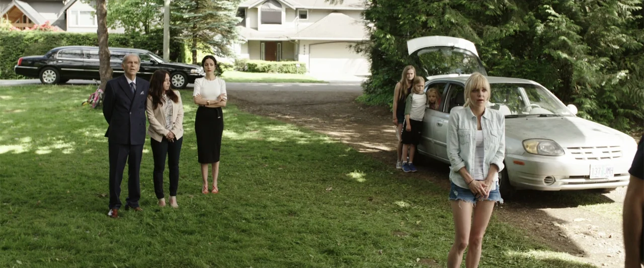 Overboard (2018) movie still: fullbody — A suburban front yard features a group of people standing on green grass. A woman in a denim shirt faces…