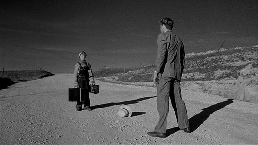 Paper Moon (1971) movie still: wide — A desolate dirt road stretches through an open landscape with sparse vegetation. A man in a suit faces a small…