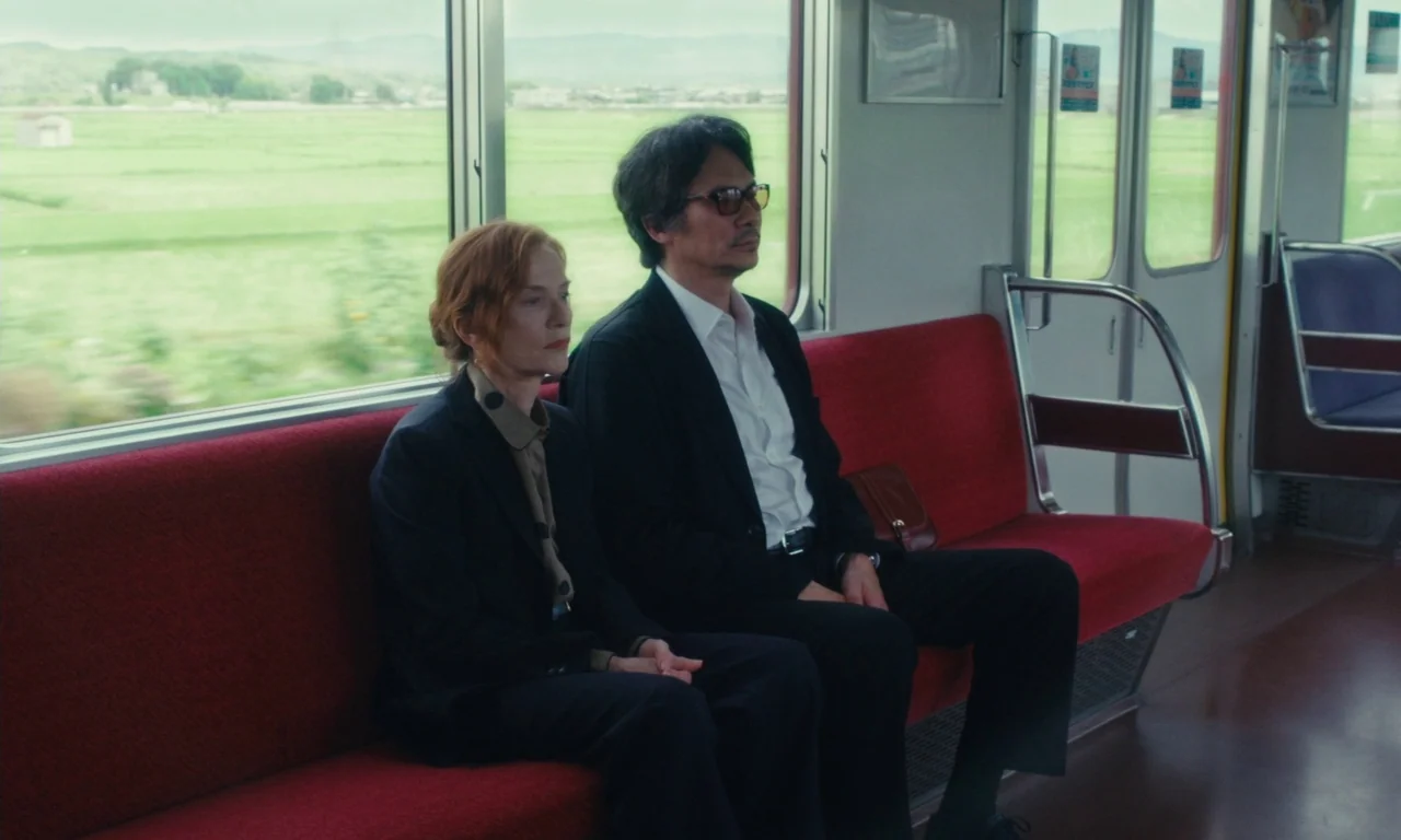 Sidonie in Japan (2023) movie still: medium — Two individuals sit side-by-side on the red bench of a sparsely occupied train. Green fields pass…