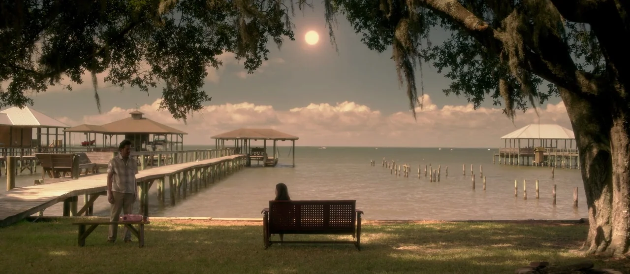 Gerald’s Game (2017) movie still: wide — A grassy waterfront area features wooden piers extending into a calm body of water. A man stands beside a…