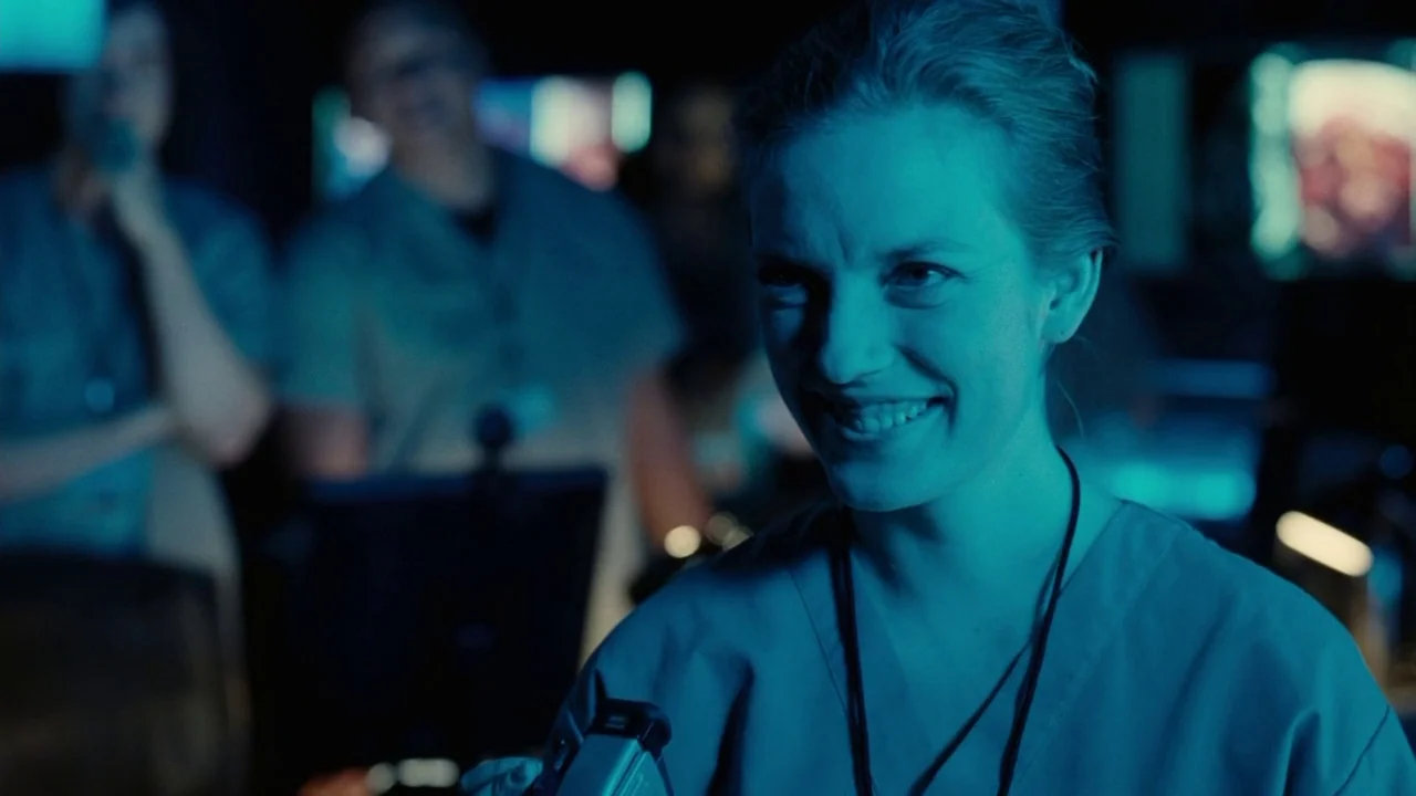Splice (2009) movie still: closeup — A dimly lit room filled with medical monitors and glowing display screens. A woman wearing scrubs smiles…