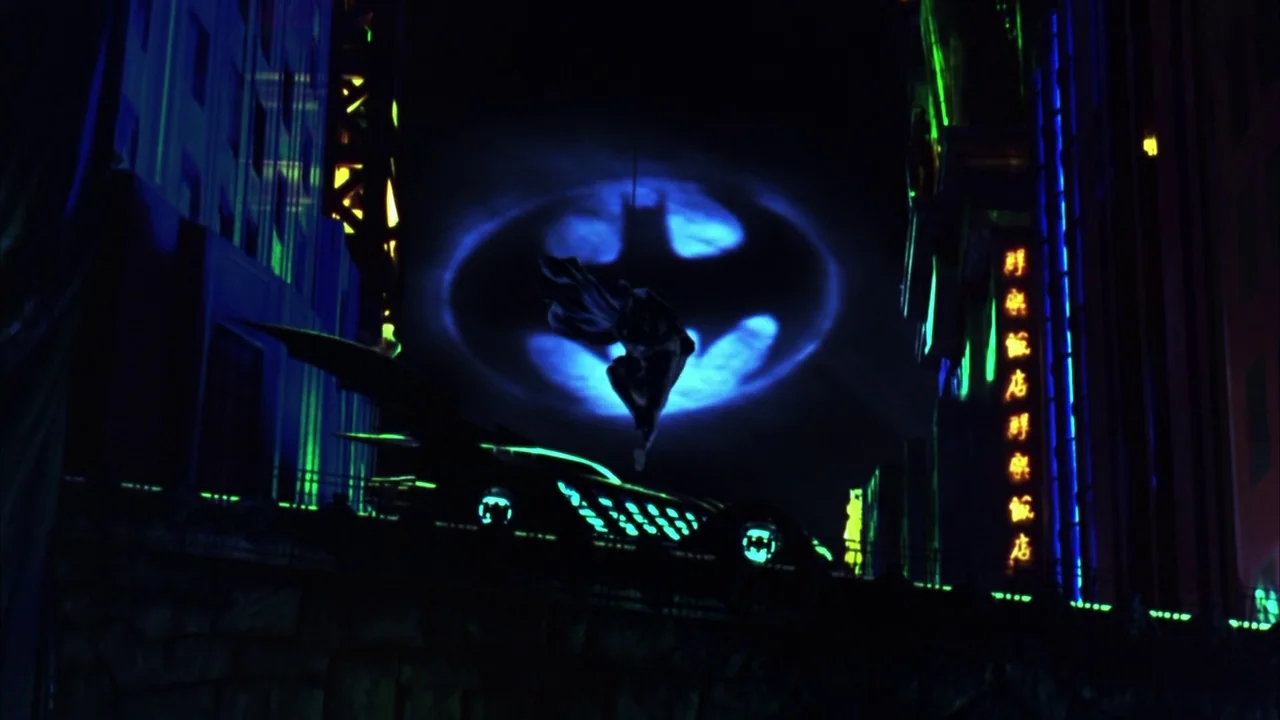 Batman Forever (1995) movie still: wide — A dark urban environment is filled with vertical buildings and vibrant green neon signs. A costumed hero…