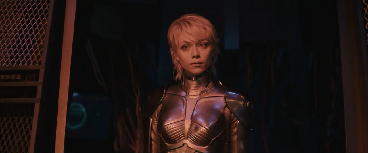 M3GAN 2.0 (2025) movie still: medium — A dark environment features a metallic-clad figure standing in a dimly lit passageway. The subject, a woman…