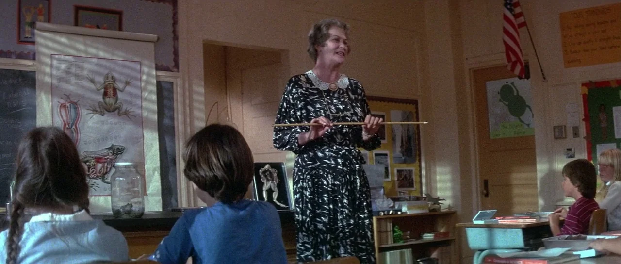Invaders From Mars (1986) movie still: medium — An elementary science room features anatomical frog posters and student desks in rows. A teacher in…