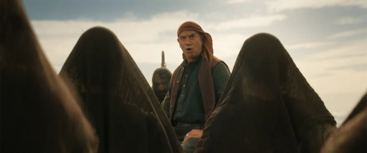 The Bluff (2026) movie still: medium — An arid, open landscape under a hazy daylight sky. A middle-aged man stands facing a group of individuals…