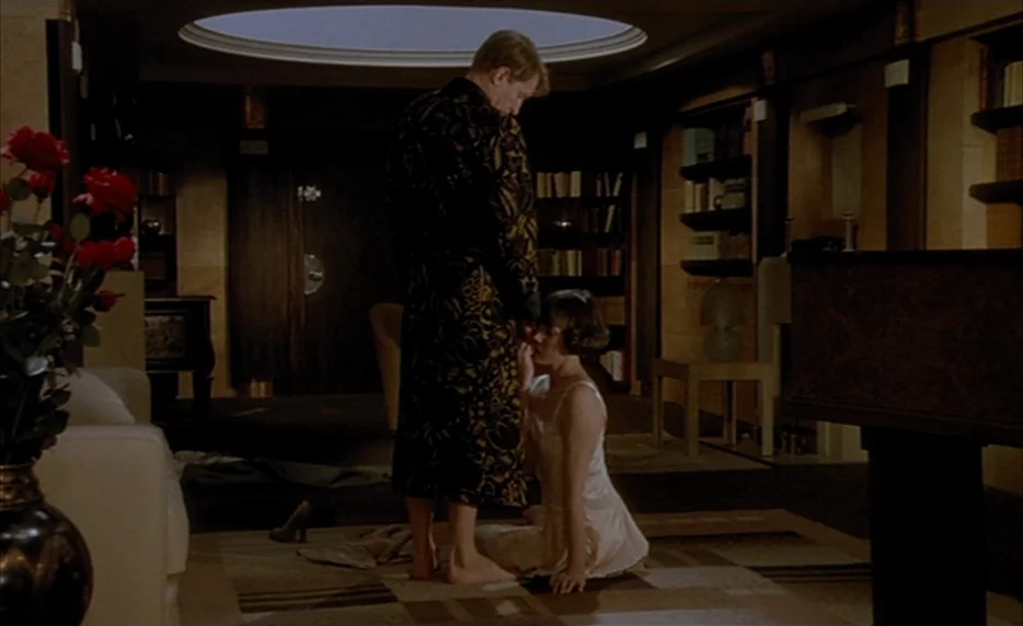Mélo (1986) movie still: fullbody — A dimly lit room with art deco architectural details and built-in bookshelves. A man stands wearing a robe while…
