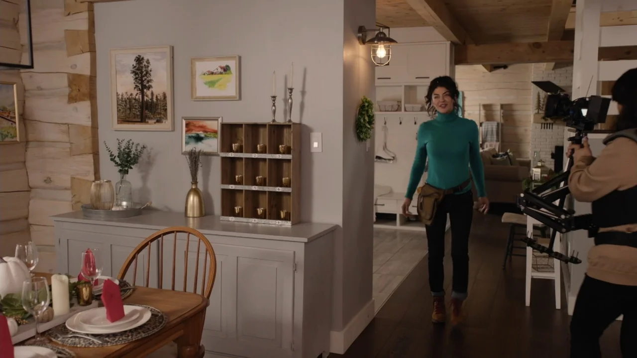 Designing Christmas (2022) movie still: fullbody — A modern rustic interior space featuring log walls and a formal dining table. A woman walks…