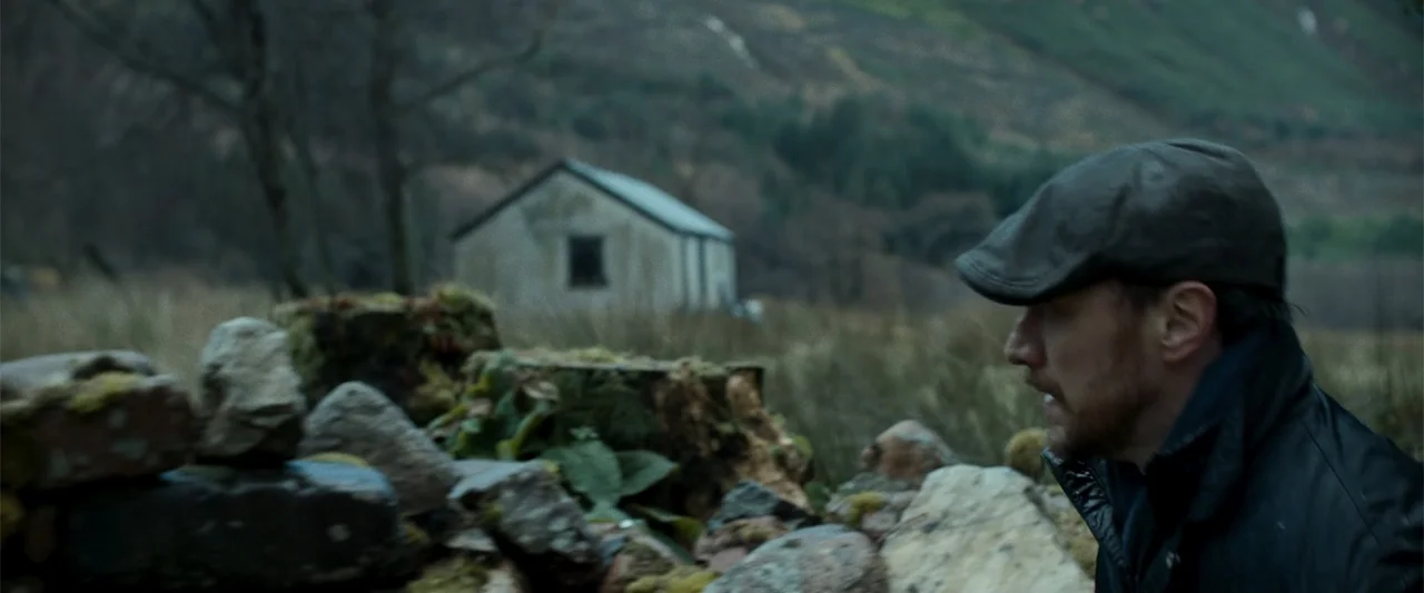 My Son (2021) movie still: medium — A desolate Highland landscape features a stone wall and a distant small cottage. A man crouches behind the stone…