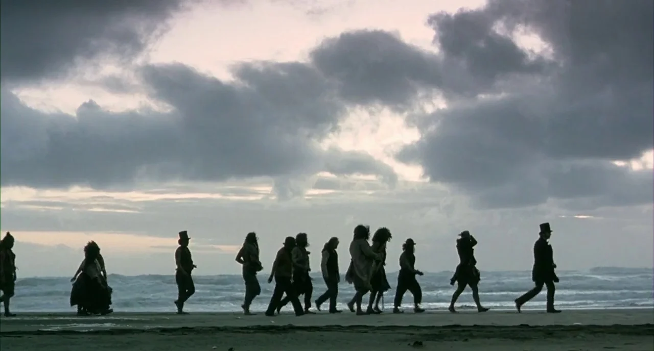 The Piano (1993) movie still: wide — A group of figures in 19th-century attire walk along a wet sandy beach. Dark storm clouds fill the vast sky…