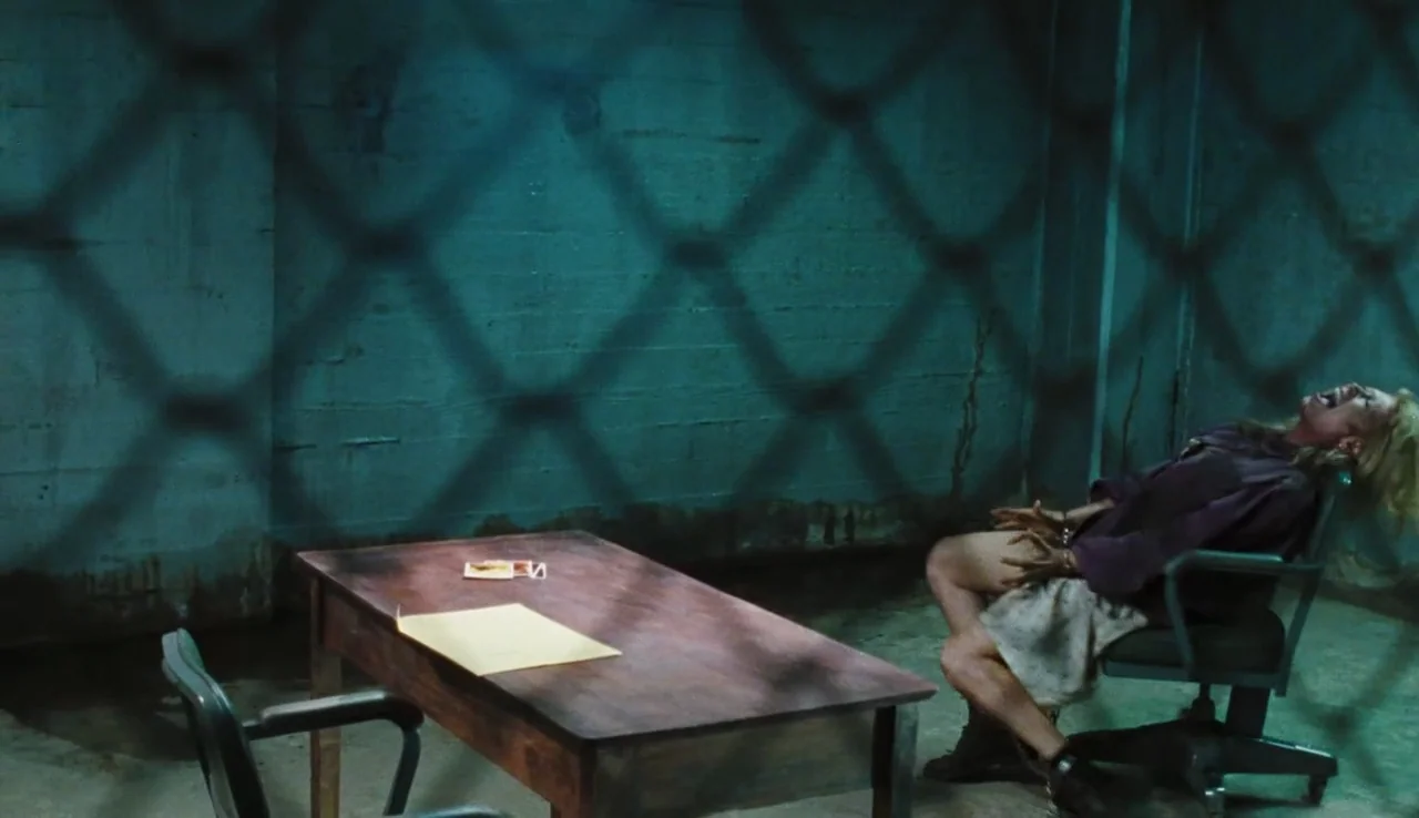 The Devil’s Rejects (2005) movie still: wide — A dimly lit room features a concrete wall and a lone wooden table. A woman in a chair is chained and…