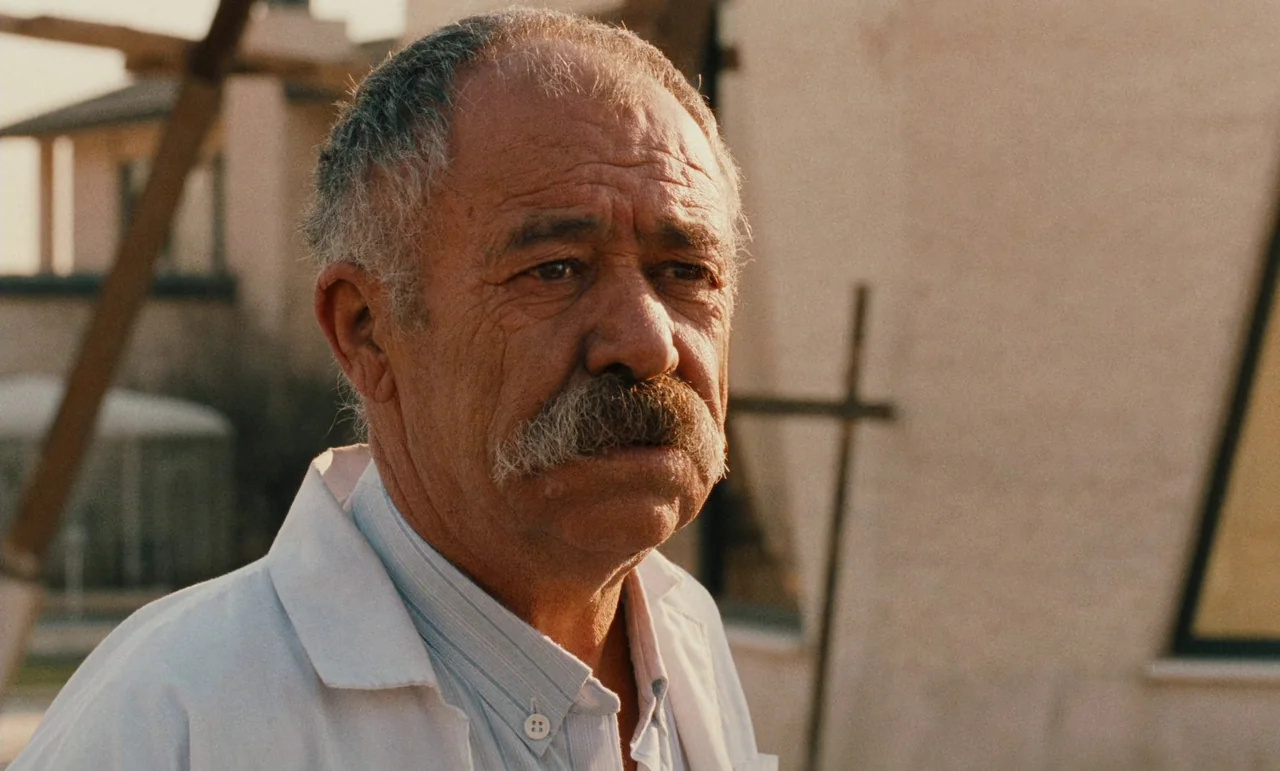 Taste of Cherry (1997) movie still: closeup — An older man stands outdoors in front of a light-colored stucco building structure. He wears a lab…