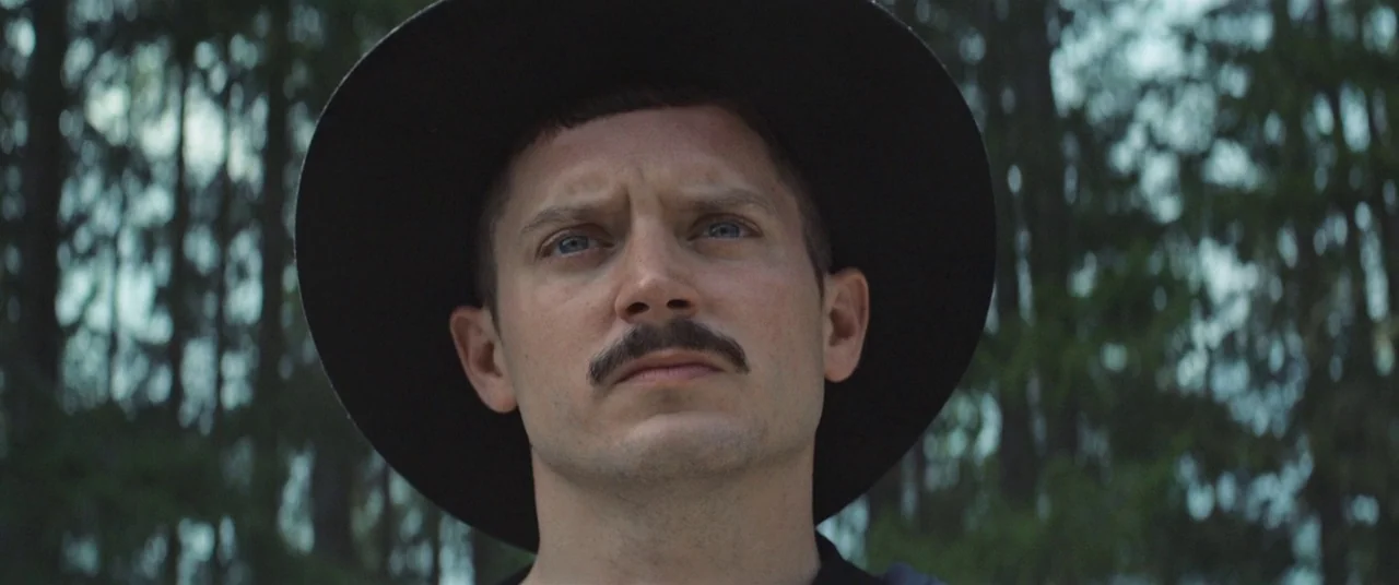 Come To Daddy (2019) movie still: closeup — A dense forest with blurred tree trunks serves as the background. A man wears a black wide-brimmed hat…