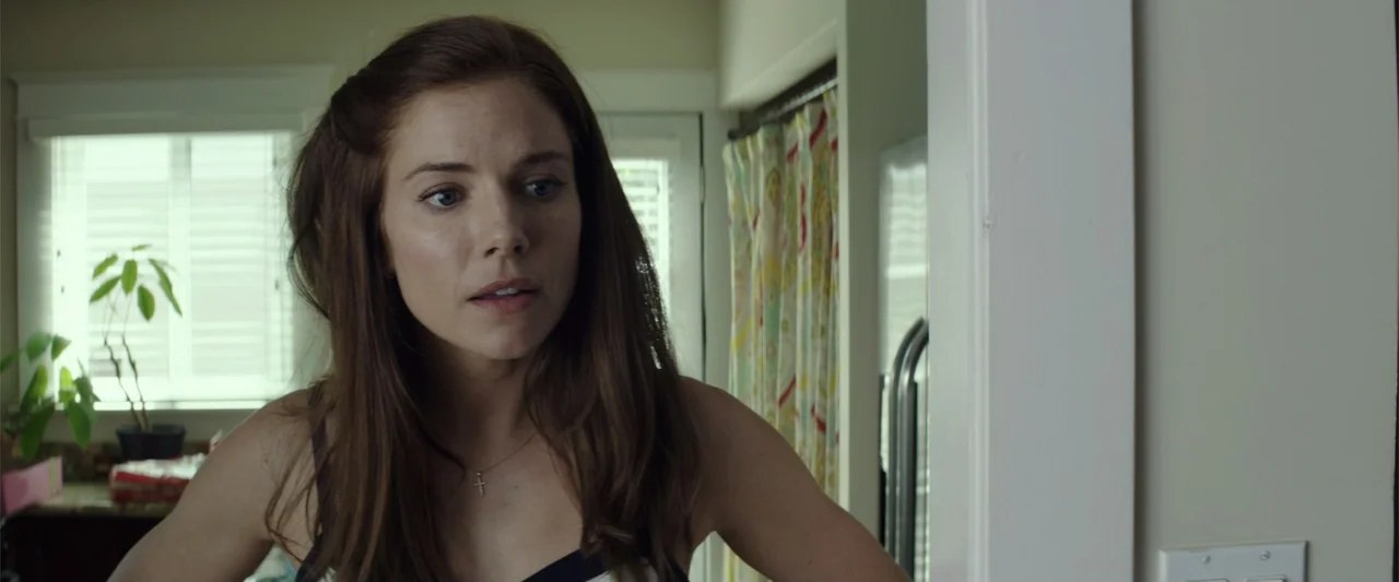 American Sniper (2014) movie still: medium — A residential room features a white door frame, a potted plant, and patterned curtains. A woman in a…
