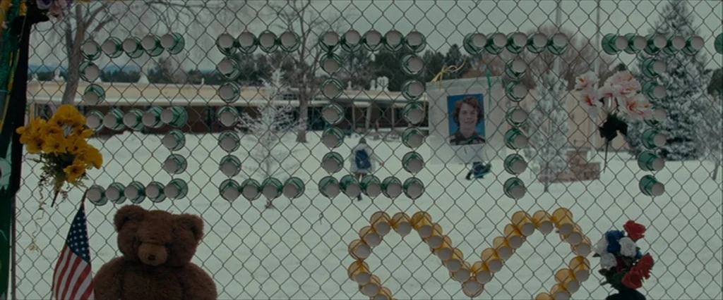 Let Me In (2010) movie still: wide — A chain-link fence holds a memorial with plastic cups, a photograph, and teddy bear. The fence stands in front…
