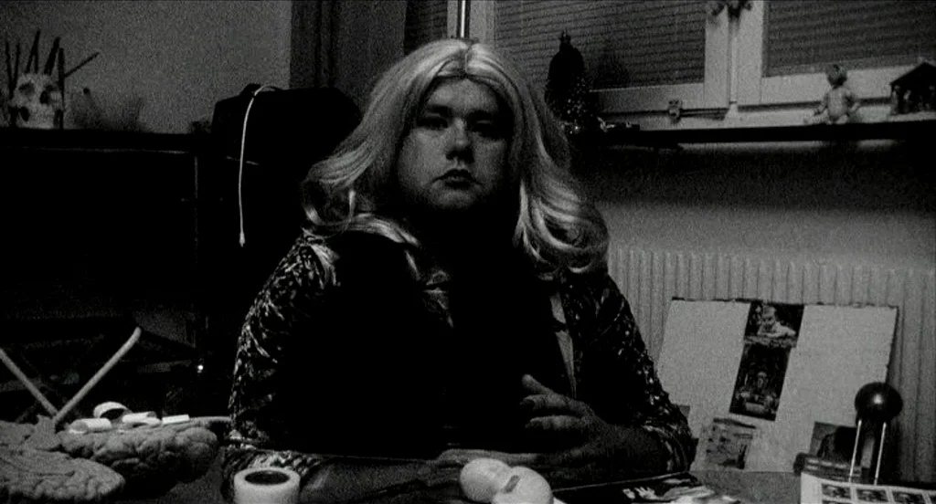 Container (2006) movie still: medium — A dark indoor space featuring a desk cluttered with various small objects and papers. A person wearing a…