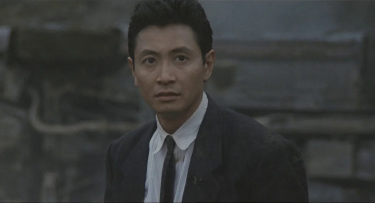 Godzilla vs. Biollante (1989) movie still: medium — An urban outdoor setting features a blurred background of gray textures. A man stands in the…