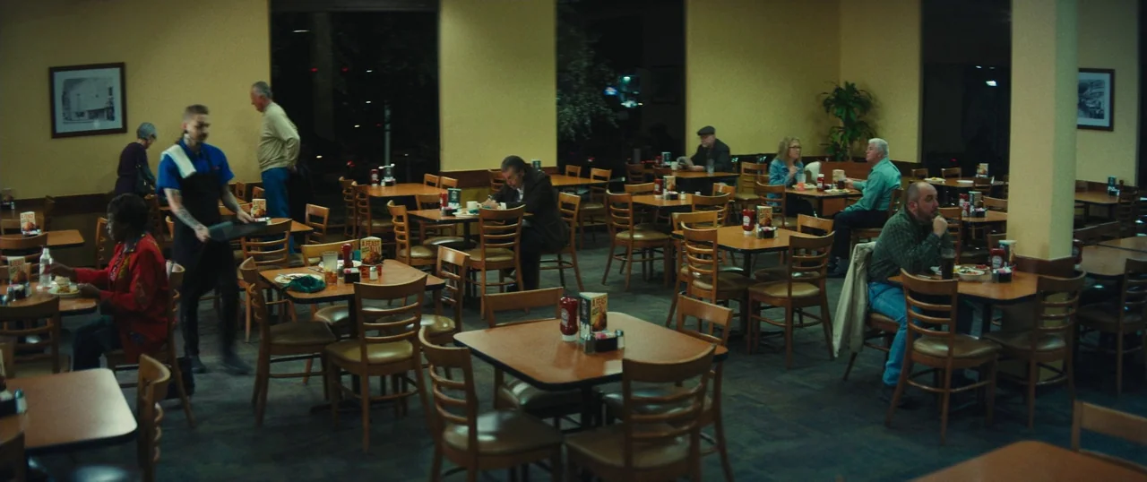 Manglehorn (2014) movie still: wide — A quiet dining area with rows of square tables and wooden chairs at night. Several patrons sit at different…