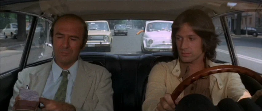 Four Flies On Grey Velvet (1971) movie still: medium — The interior of a moving car shows a driver and passenger on a city street. Several vintage…