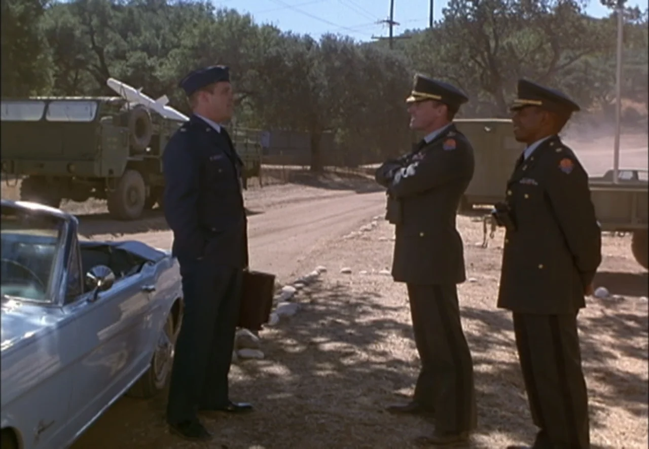 The Pentagon Wars (1998) movie still: fullbody — A dry, dusty military compound features a parked vintage convertible and tactical trucks. Three men…