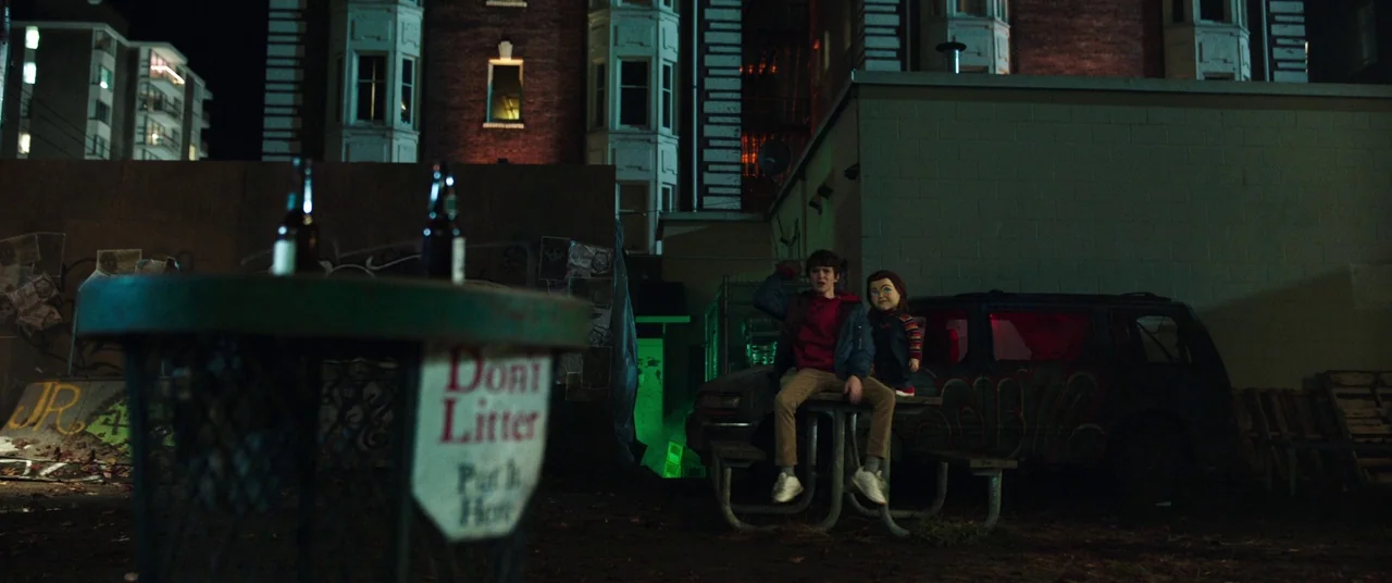 Child's Play (2019) movie still: wide — A dark night-time parking lot behind a multi-story brick apartment building with graffiti. A teenager and a…