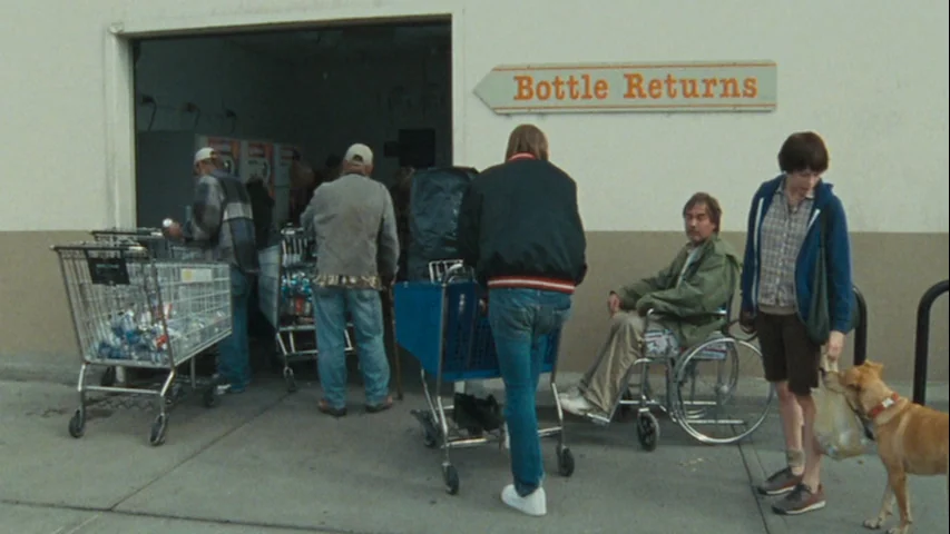 Wendy and Lucy (2008) movie still: wide — A group of people waits at the entrance of a concrete block building. They have several shopping carts…