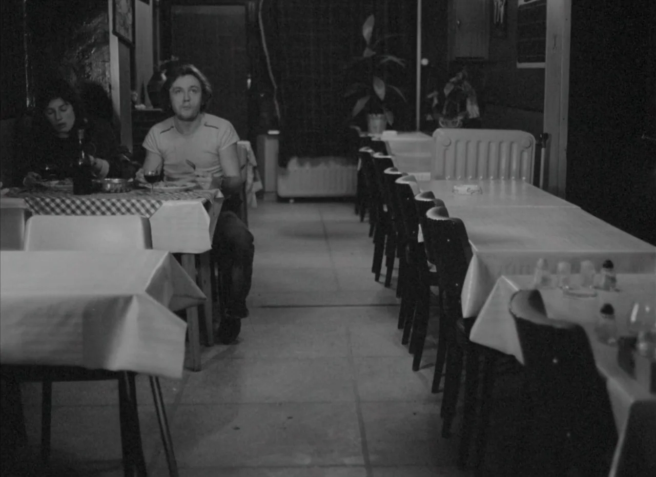 Je Tu Il Elle (1974) movie still: medium — A quiet restaurant interior features rows of tables covered in white cloths. A man and a woman sit…