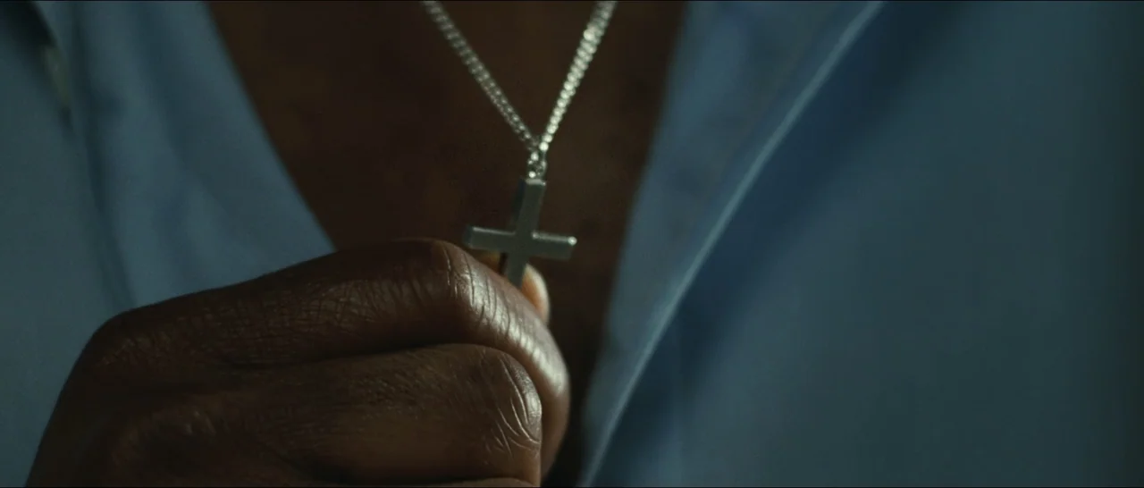 Slackers (2002) movie still: closeup — A close-up view shows a hand holding a small metal crucifix pendant. The background reveals a blurred portion…
