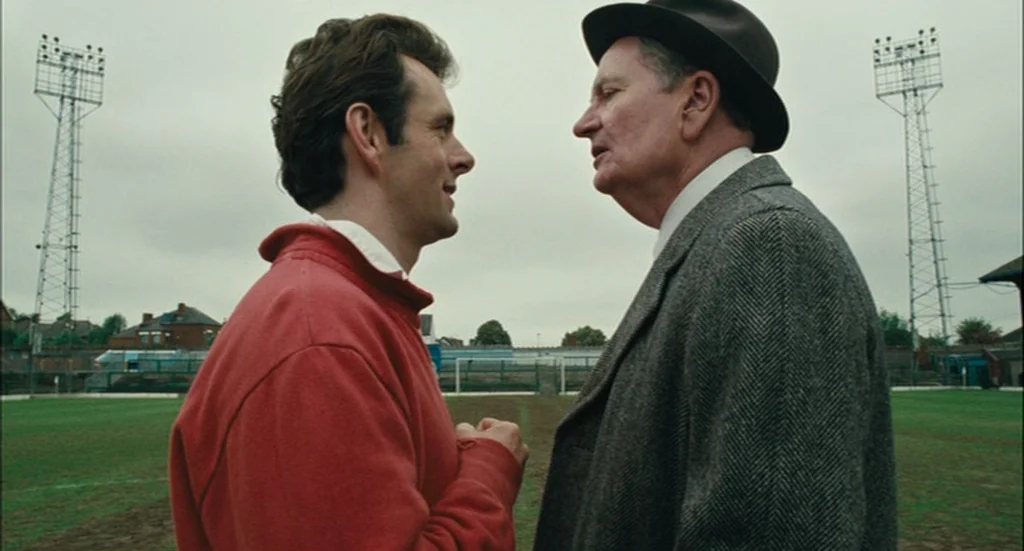The Damned United (2009) movie still: medium — An outdoor soccer pitch with tall metal floodlight towers on an overcast day. Two men stand facing…