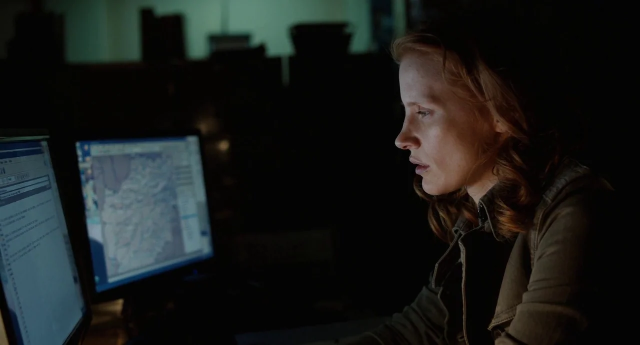 Zero Dark Thirty (2012) movie still: closeup — A darkened, sparse room is lit only by the screens of two computer monitors. A woman in a green…