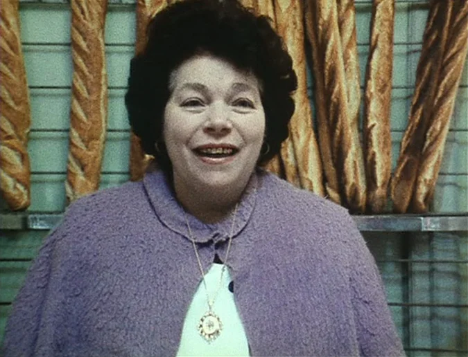 Daguerréotypes (1976) movie still: medium — A woman stands in front of a rack of baguette loaves at a bakery. She is facing forward while smiling at…