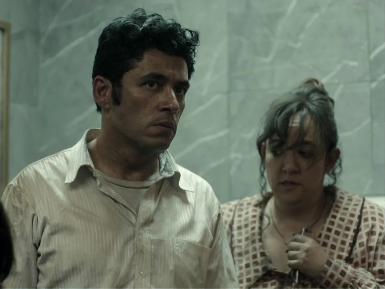 November (2025) movie still: medium — A cramped bathroom features light grey veined tiles on the walls. A man in a striped shirt and a woman in a…