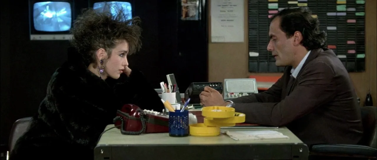 Subway (1985) movie still: medium — A small security room features black wall monitors, a desk, and organizational charts. A woman with voluminous…