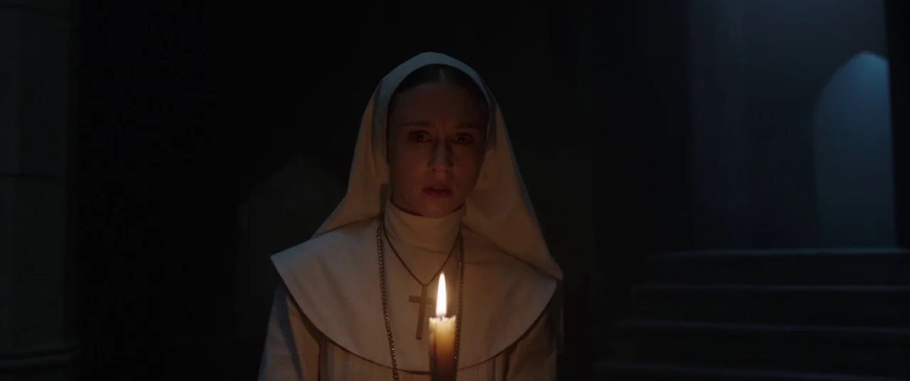 The Nun (2018) movie still: medium — A dark, stone-walled corridor is illuminated only by a single small flickering candle flame. A young nun stands…