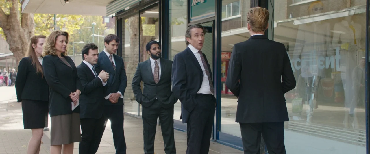 Greed (2019) movie still: fullbody — A group of men and women stand on a paved walkway outside a storefront. The group faces a man standing near a…