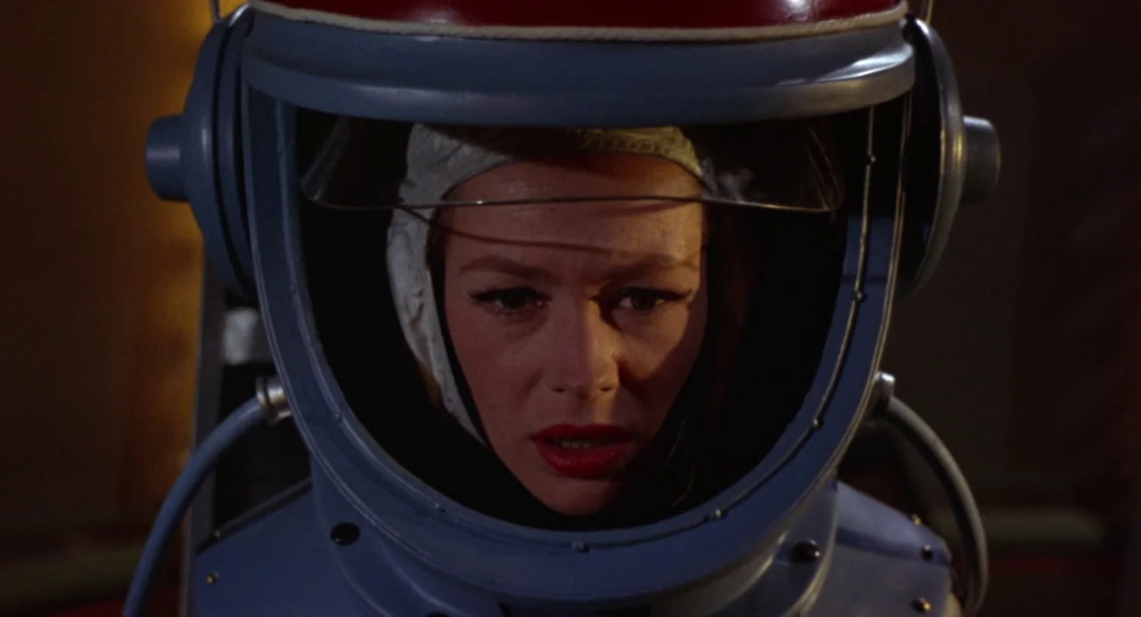 Queen of Blood (1966) movie still: closeup — A woman wears a large blue space helmet in a dark environment. The helmet has a white cap visible…