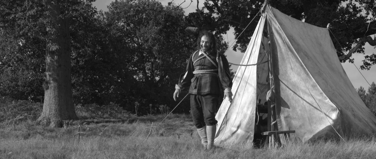 A Field In England (2013) movie still: fullbody — A grassy field features a large, worn canvas tent situated near several large trees. A man with a…