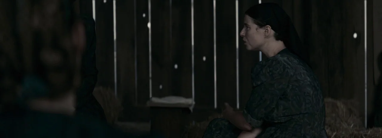 Women Talking (2022) movie still: medium — The interior of a rustic barn features dark vertical wooden boards with sunlight slits. A woman in a dark…