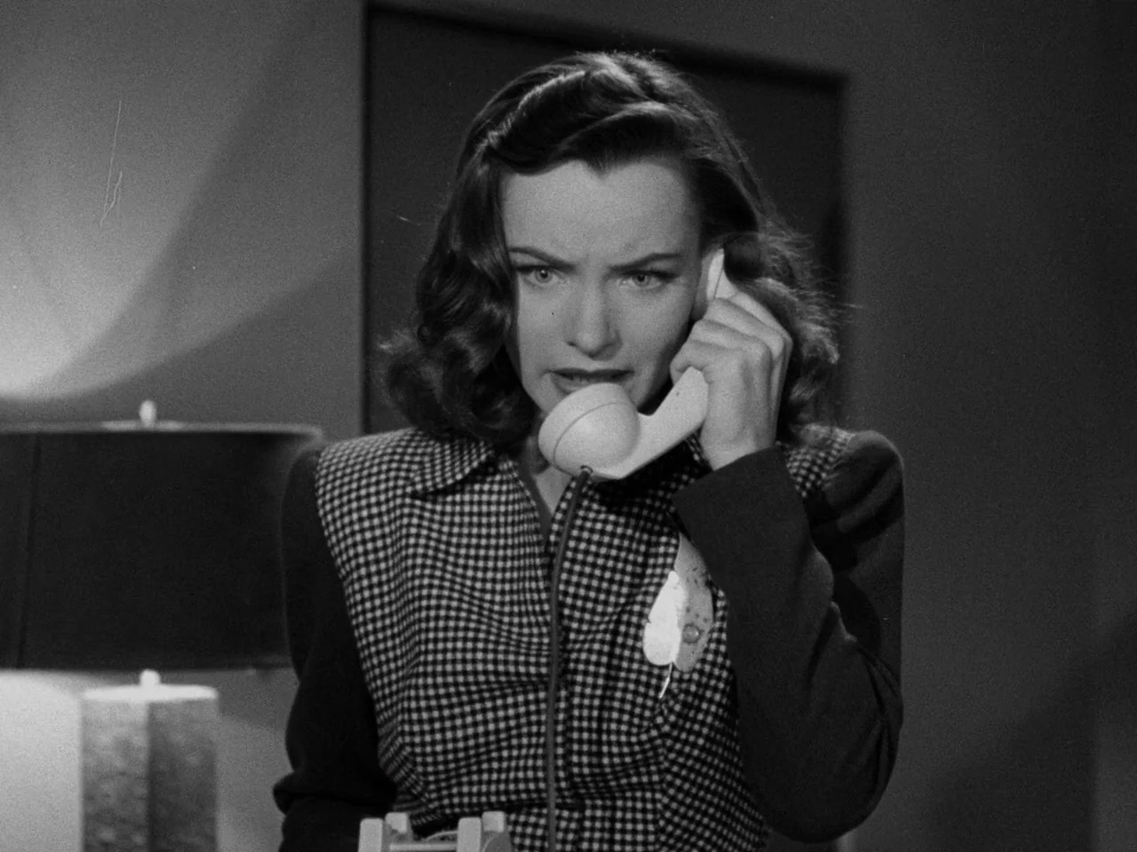 Phantom Lady (1944) movie still: medium — A woman stands in a dimly lit room with a lamp visible to the side. She holds a vintage telephone receiver…