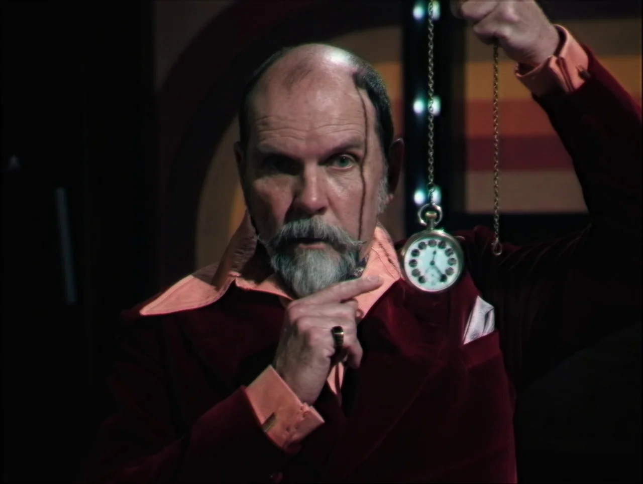 Late Night With The Devil (2023) movie still: closeup — A man in formal velvet attire stands against a dark, indistinct background. He points his…