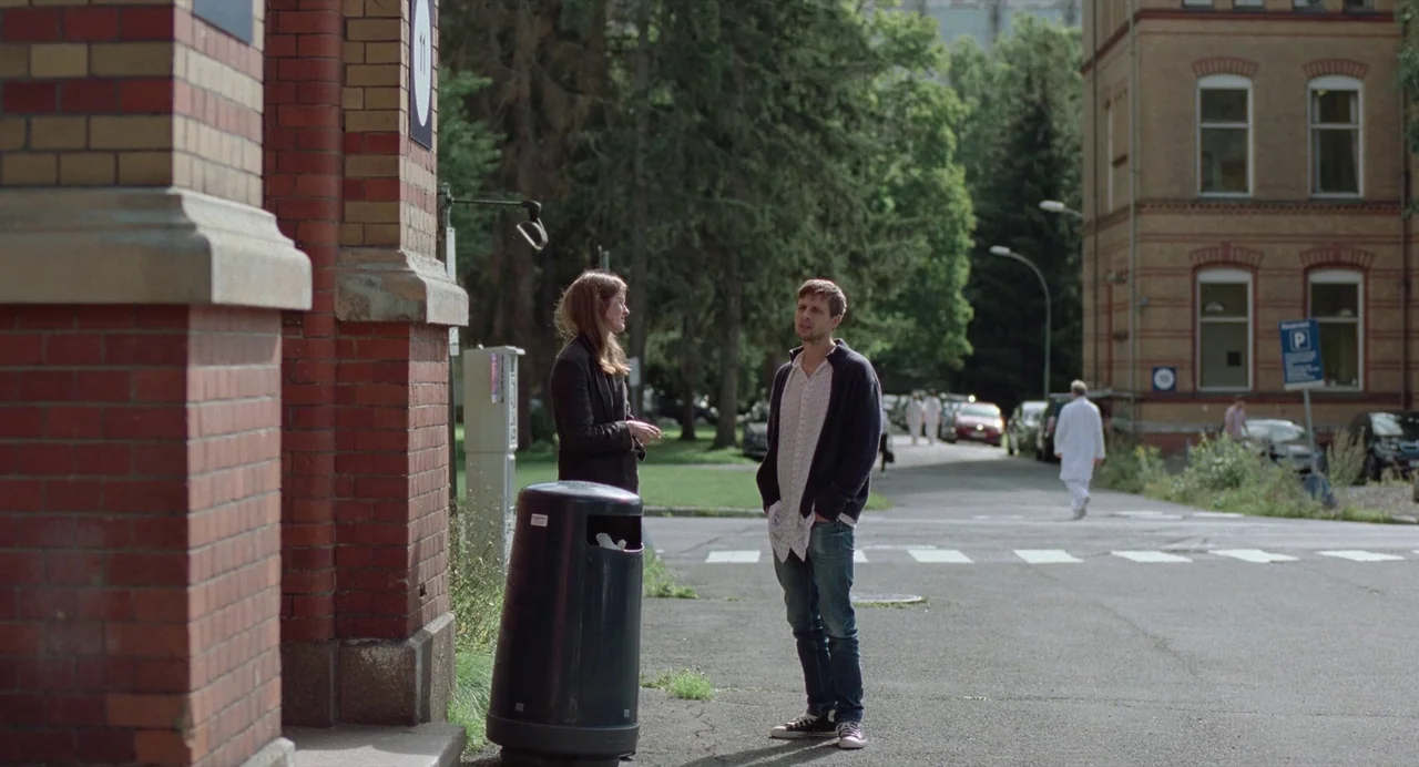 The Worst Person in the World (2021) movie still: wide — A paved path between red brick hospital buildings surrounded by mature green trees. Two…