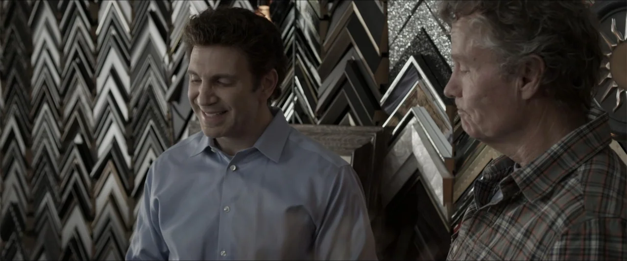 Heavenly Deposit (2019) movie still: medium — A frame shop interior filled with numerous picture frame samples mounted on the wall. Two men stand…