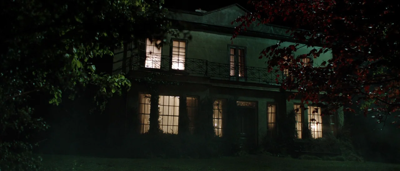 The Hallow (2015) movie still: establishing — A large two-story house stands at night behind dark tree branches and foliage. The windows are…