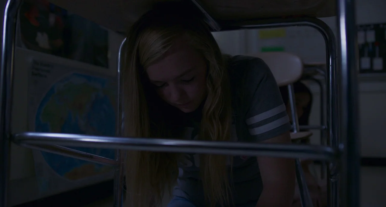 Eighth Grade (2018) movie still: medium — A dimly lit classroom interior features metal desk frames and educational posters on walls. A teenage girl…